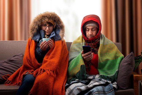 Middle age family in parkas drinking coffee on the sofa while using smartphone in their cold living
