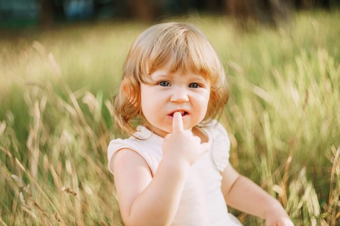 Little girl sits outdoors and holds a finger in his mouth.