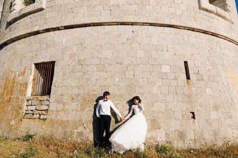 Bride and groom are holding hands and standing tjgether near the wall of the Arza fortress.