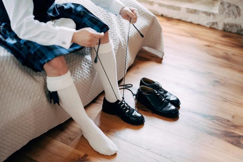 Preparing for a Scottish wedding. Man in a kilt, sporran and high socks sits on the bed and tie