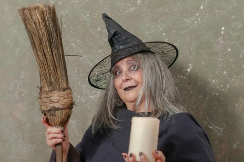 Old witch with white hair and a black hat holding a broom in her hand and a candle in the other