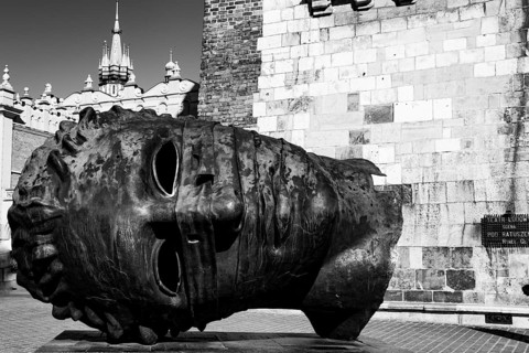 A large metal head sculpture lying on its side near a historical building in black and white,