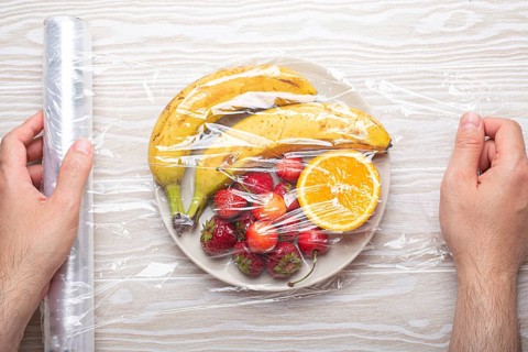 Male hands wrapping assorted different fruit and berries on white plate in plastic clear film top
