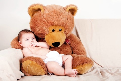 Cute Caucasian Hispanic unisex baby in the arms of a big brown stuffed teddy bear on the couch, New