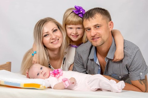 Young parents and daughter sit at the five-year crib, the crib is on a two-month baby on her back