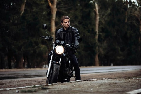 A person in a leather jacket sits on a motorcycle by a forest road, creating a moody atmosphere