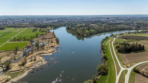 View of a river (Danube) with riverside vegetation, neighbouring fields and a town in the distance,