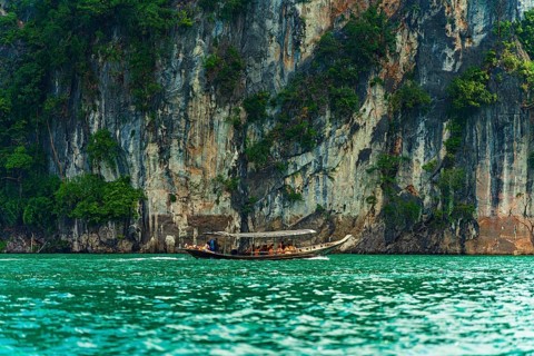 Longboat navigating along a towering cliff with greenery above turquoise water, Cheow Lan Lake,