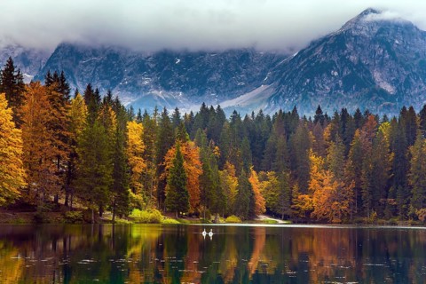 The Dolomites are covered with clouds. Orange and yellow trees are reflected in the green smooth