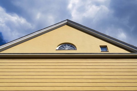 A yellow house with a pitched roof and one semi-circular and one square window in front of a cloudy