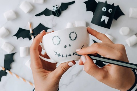 Female hands paint on white cup scary jacks face DIY for kids Halloween home activities Holiday art