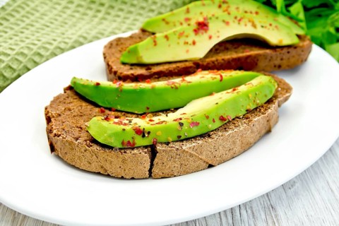 Two slices of rye bread with slices of avocado and pepper on a plate, green cloth, parsley on the