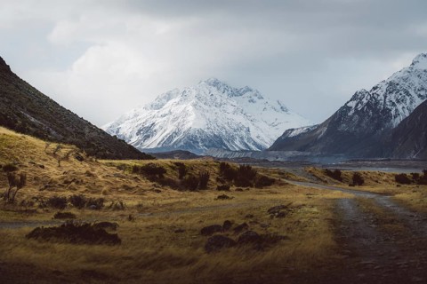Gravel track leads through a vast landscape with snow-capped mountains and cloudy skies, Hooker