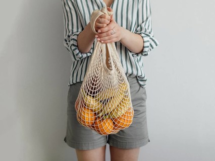Mesh bag with fruits in female hands. Stylish young woman hold mesh shopping bag on light gray wall