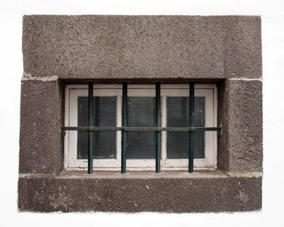 A small white wooden window with glass panes covered by steel bars in a large grey stone frame