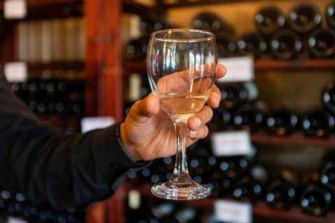 A glass of white wine on a man hand selective focus close up view against wine bottles on storage