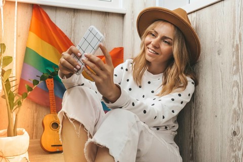 Young millennial hippie woman sitting on balcony using mobile phone. LGBTQ rainbow flag on