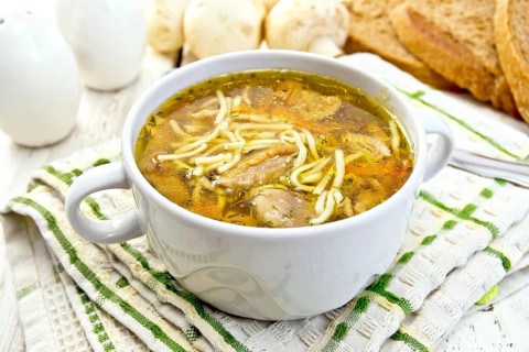 Soup with noodles and mushrooms in a white bowl on a napkin, bread on a wooden boards background