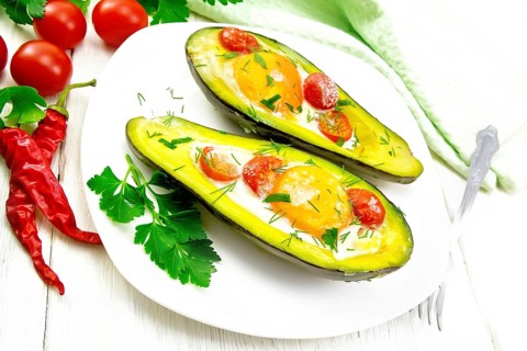 Scrambled eggs with cherry tomatoes in two halves of avocado in a plate, towel and fork on white