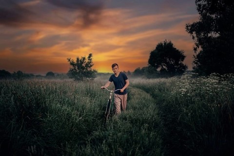 Young man walks with a retro bike along a field road, vintage style in the village at sunset.