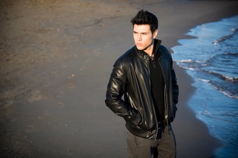 Handsome fashionable young man at the seaside on the beach with his black leather jacket looking