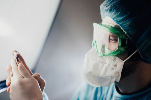 A doctor in green goggles examines a vial while wearing a mask