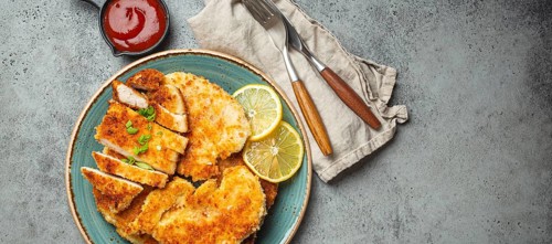 Crispy panko breaded fried chicken fillet with green salad and lemon cut on plate on gray