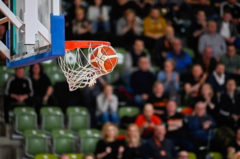 Ball falls in the basket during basketball match. Audience in the background.