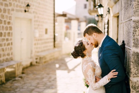 Groom kisses bride near the gray stone wall of the old house.