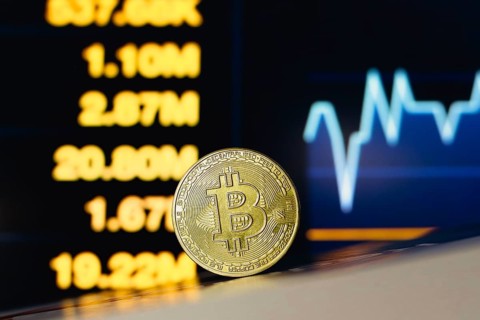 In this photo illustration, a Bitcoin coin is seen on the table and in the background, a stock