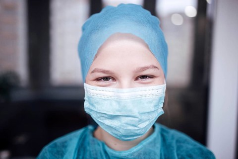 A person in blue scrubs and a medical mask smiles indoors, indicating a healthcare setting