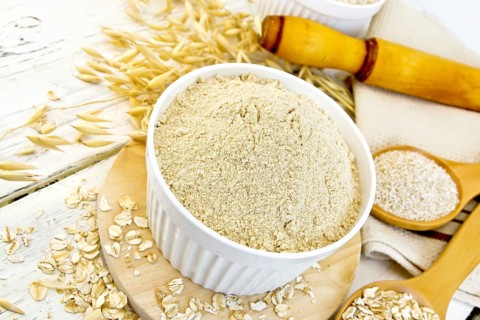 Oat flour in white bowl, oatmeal and bran in wooden spoon, stalks of oats, a rolling pin and a