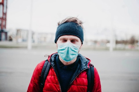 Man in a red jacket and face mask outdoors, with city buildings in the background