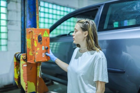A woman in a workshop thoughtfully examines a switch on a car lift, partially obscured by the car