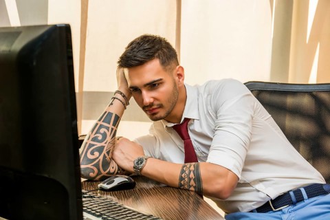 Tired bored young businessman sitting at his desk in front of his computer yawning, holding his