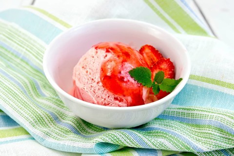 Strawberry ice cream in a white bowl with strawberries and strawberry syrup on a striped linen
