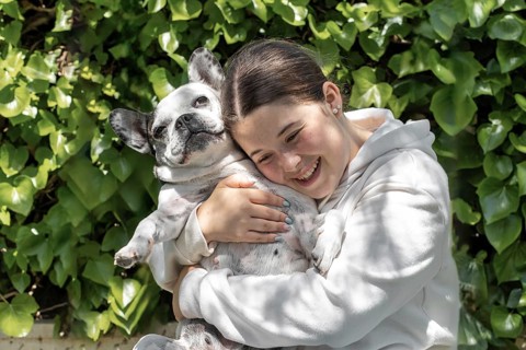 A girl in a white sweatshirt smiling and hugging a French Bulldog in a garden with lush green