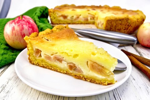 Piece of apple pie with sour cream sauce in a white plate, cinnamon, fruit and napkin on a wooden
