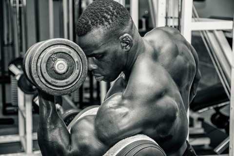 Hunky muscular black bodybuilder working out in gym, exercising biceps with dumbbell. Black and