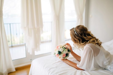 A bride in a gentle peignoir with a bouquet in her hands laying on the bed by the window with white