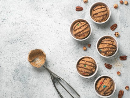 Safe-to-eat raw monster cookie dough in small portion bowl, ice cream scoop and nuts on gray cement