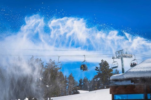 Snow cannon spreading fresh snow powder on a ski slope. Skiers riding up on chair lift. Winter ski