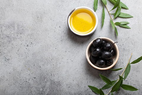 Black olives and extra virgin olive oil in little bowl with olive tree branches top view on gray