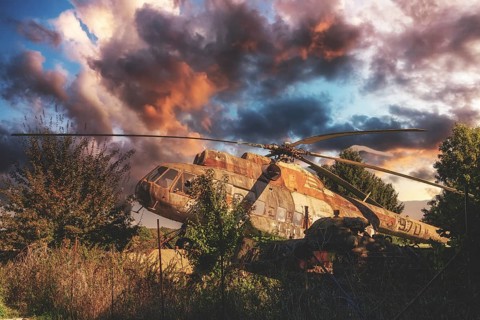 An abandoned, rusty helicopter stands overgrown in nature under a dramatic, cloudy sky at sunset,