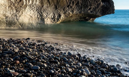 Low angle view of seascape with windy waves splashing on the rock and the sea pebbles. Rock of