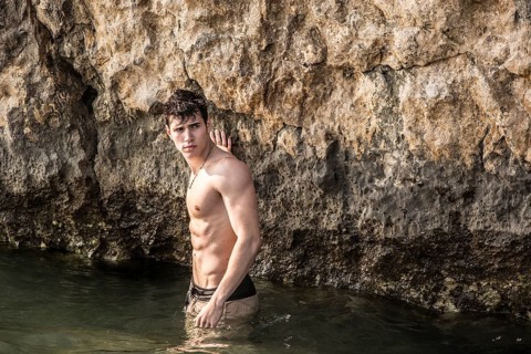 Attractive young shirtless athletic man standing in water by sea or ocean shore, wearing shorts,