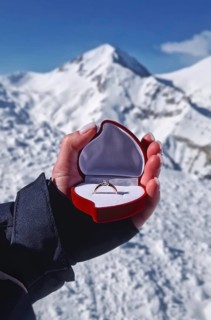 Female hand closeup holding an engagement ring inside a red heart shaped box over the snowy