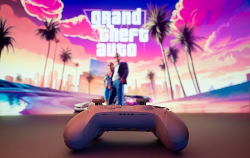 A close-up of a white gamepad with the Grand Theft Auto VI seen on screen in the background. GTA VI