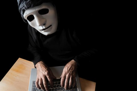 Hacker dressed in black with mask and hood typing on a computer while looking at the camera