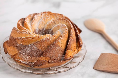 Fresh marble bundt cake, cake with icing sugar next to a rustic wooden spoon on a light-coloured